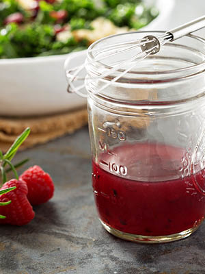 Himbeerdressing im Glas mit Schneebesen oben daruf. Salat im Teller im Hintergrund.